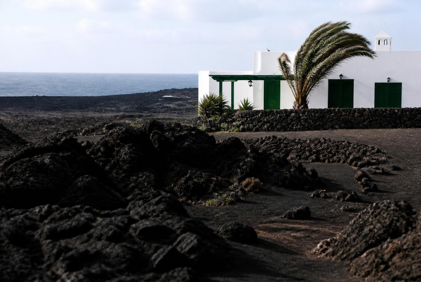 Casa blanca con carpinterías verdes junto a un campo de lava negra frente al océano en Lanzarote